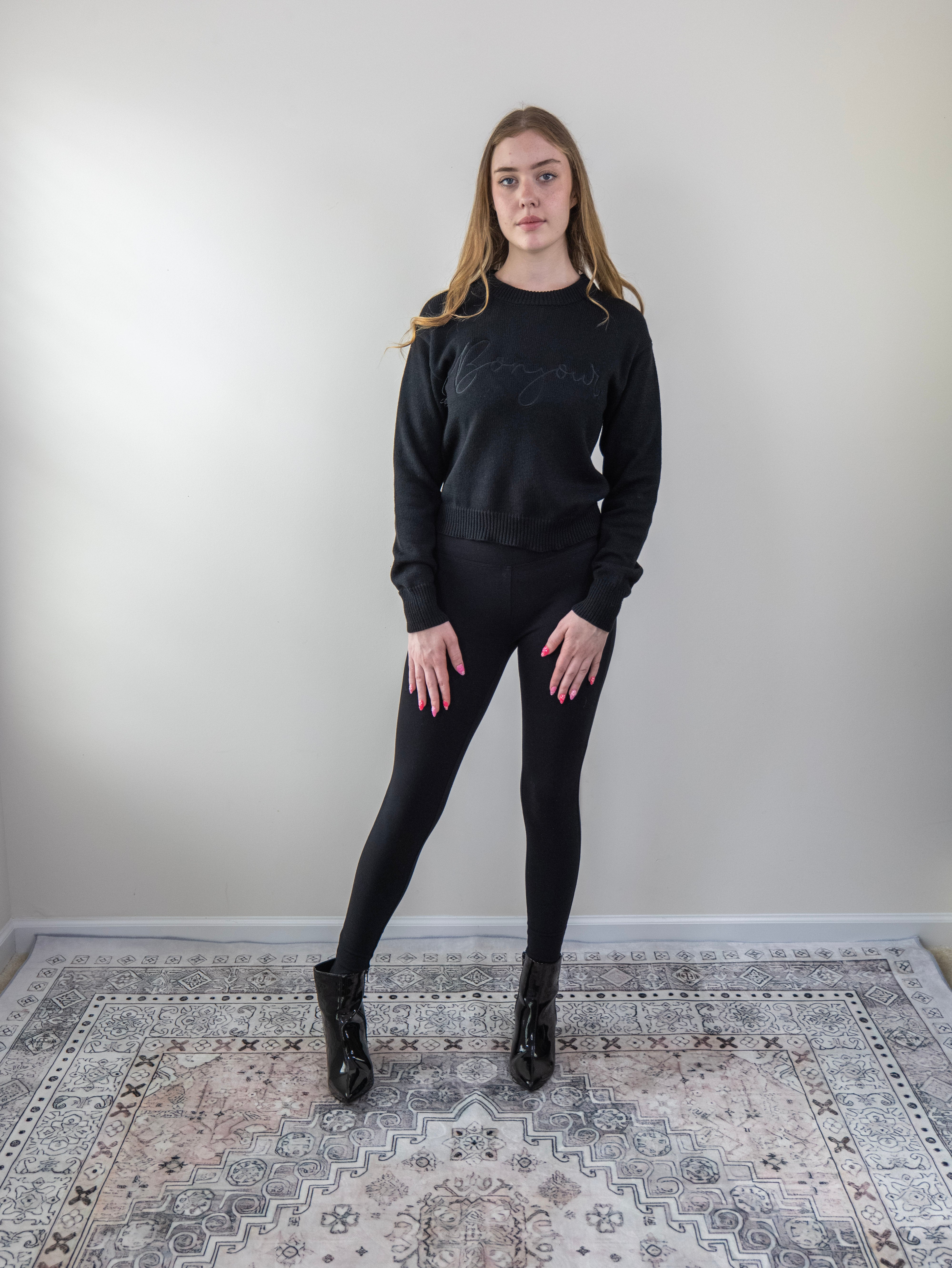 Model wearing a black “Bonjour” long-sleeve knit sweater, standing front-facing