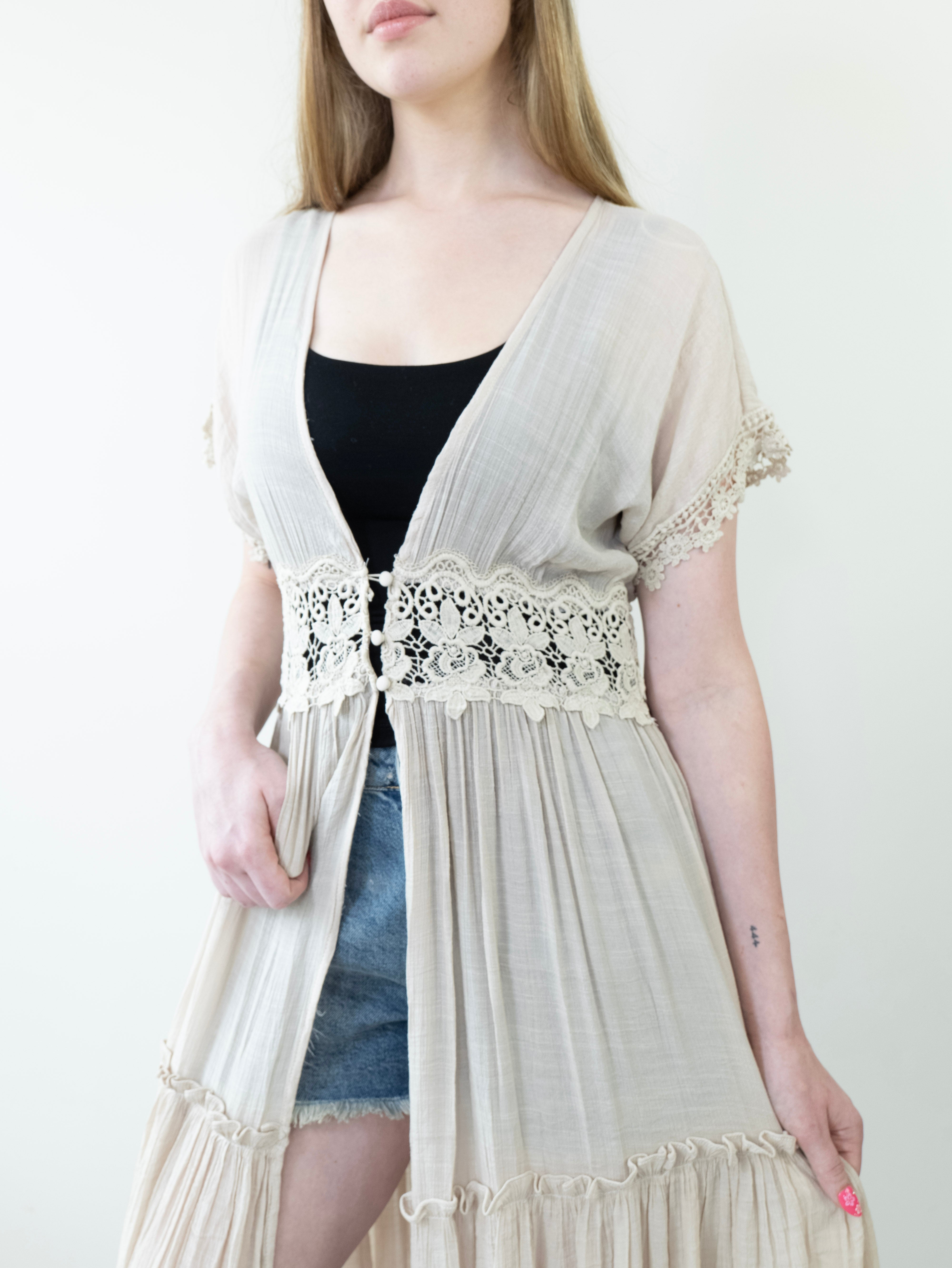 Model wearing a long beige lace-trimmed cardigan over a black top and denim shorts on a white background.