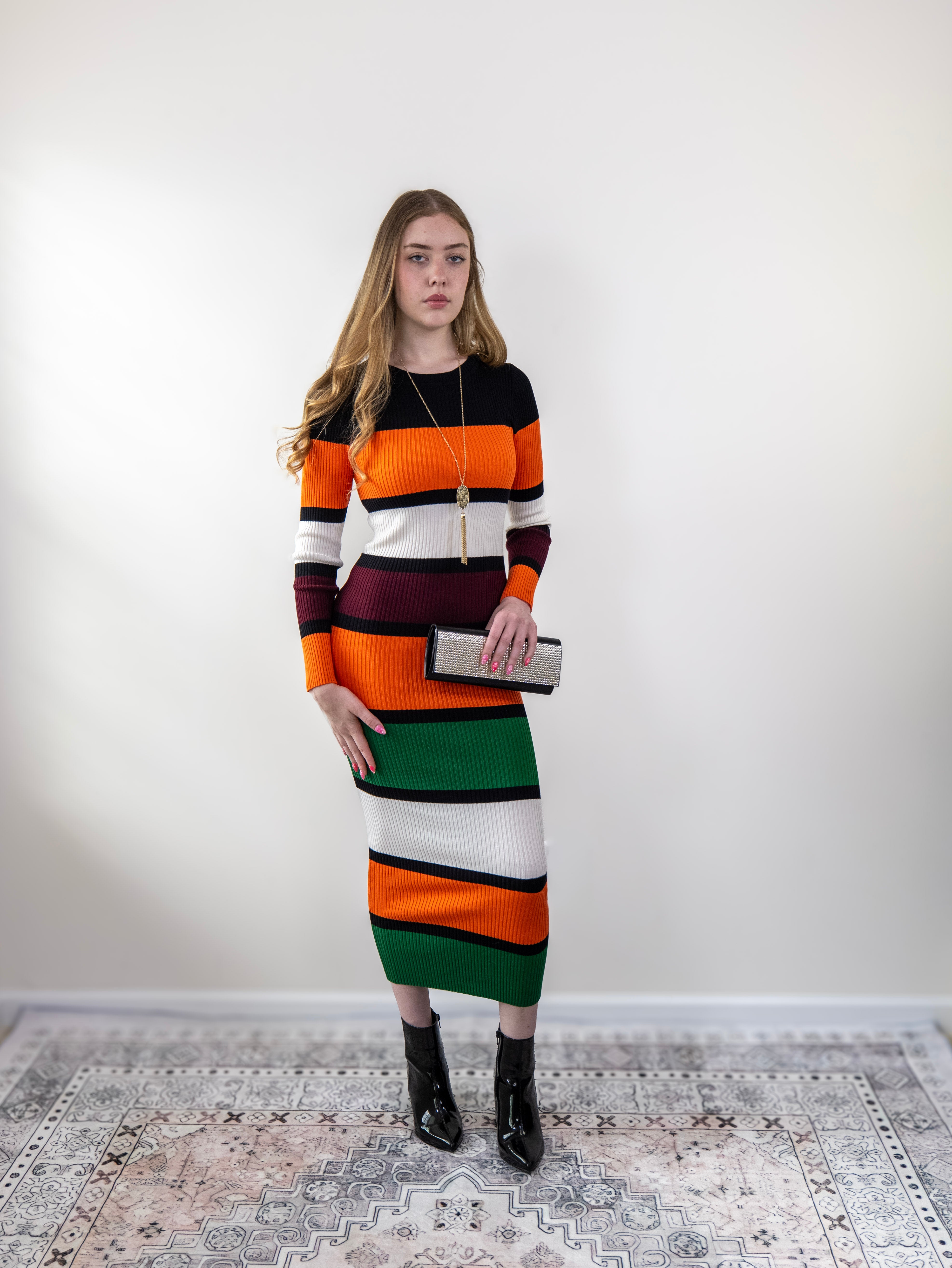 Model wearing multi-stripe orange sweater dress, hands on hips, mid-length view.